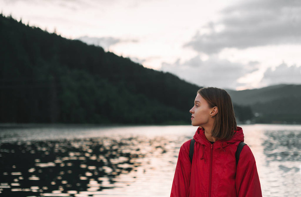 Matkustaja tyttö etsii syrjään rentouttava lähellä vuoristometsää ja järveä, nauttia kauniista näkymistä illalla. Rauhallinen muotokuva nuoresta naisesta punaisessa sadetakissa lähellä vuorilampea. Kopioi tila
. - Valokuva, kuva