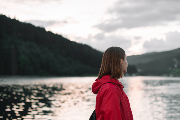 Πίσω όψη της νεαρής γυναίκας κοιτάζοντας στην άκρη απολαμβάνοντας τη θέα στη λίμνη βουνό και το δάσος. Γυναίκα τουρίστρια σε ένα κόκκινο αδιάβροχο αίσθημα χαλαρή και απρόσεκτη κοντά στη λίμνη στο ορεινό δάσος το βράδυ. - Φωτογραφία, εικόνα