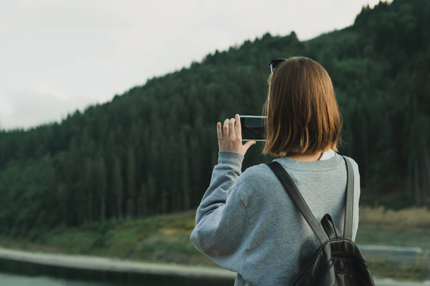 Young female traveler in grey sweatshirt taking photo on smartphone of mountain forest landscape. Back view of tourist woman using cellphone to photograph coniferous woods. Travel, vacation concept. - Photo, Image
