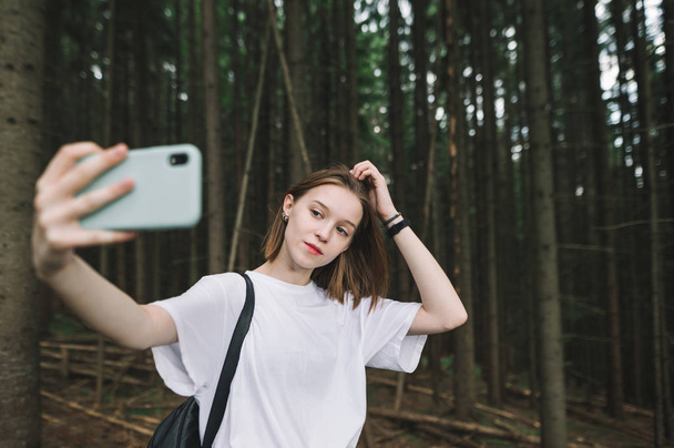 Jong schattig toeristisch meisje in het bergbos met behulp van smartphone om een selfie te nemen. Vrolijke aantrekkelijke vrouwelijke reiziger neemt selfies op haar mobiele telefoon in het naaldbos. Reisconcept. - Foto, afbeelding