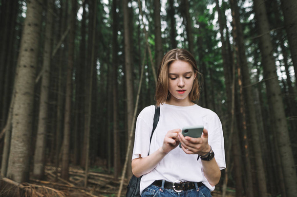 Portrait of a female tourist in the fir woods searching for the way in her phone maps application. Young beautiful traveler girl using smartphone app to look at maps and go through mountain forest. - Photo, Image