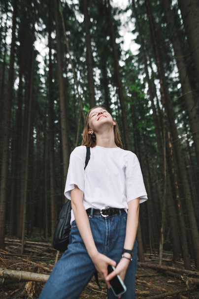 Vertikální fotografie usměvavé turistické dívky se zavřenýma očima, šťastně kráčející v jehličnatém lese. Mladá kavkazská žena cestovatel s širokým úsměvem je pozitivní a veselý v lese. - Fotografie, Obrázek