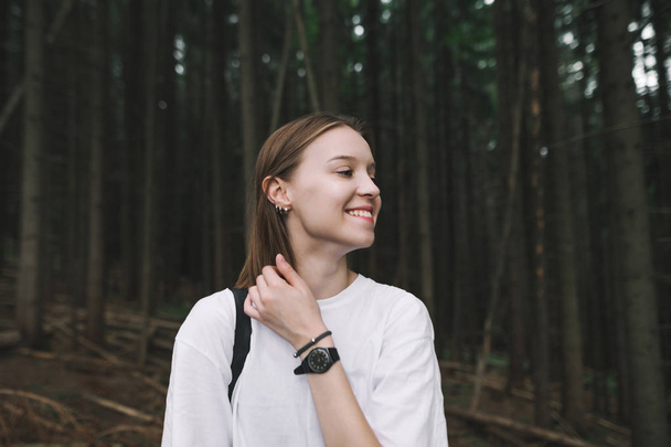 Closeup photo of a smiling young hiker woman in the fir forest feeling positive and happy. Close up shot of a girl traveler with a broad smile touching her hair looking aside in the coniferous woods. - Foto, imagen
