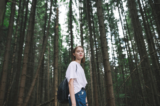 Portret uśmiechniętej kobiety z plecakiem stojącej w górskim lesie iglastym patrzącej w kamerę. Wesoła turystka stojąca wśród jodły na tle lasu. - Zdjęcie, obraz