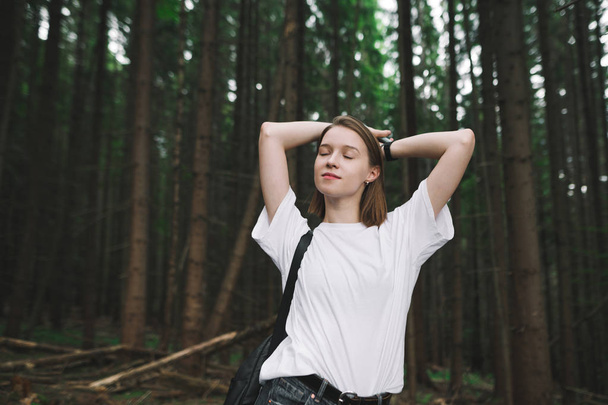 Young attractive female traveler standing in the mountain woods, hands up and behind her head, eyes closed, feeling satisfied and relaxed. Portrait of a dreamy tourist girl in the mountain fir forest. - Photo, Image