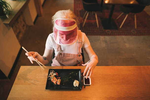Attractive girl sits in a cozy Japanese restaurant with a plate of sushi rolls and soy sauce, holds chopsticks, looks downstairs, wears a pink cap and sundress. Asian restaurant concept. Top view - Photo, Image