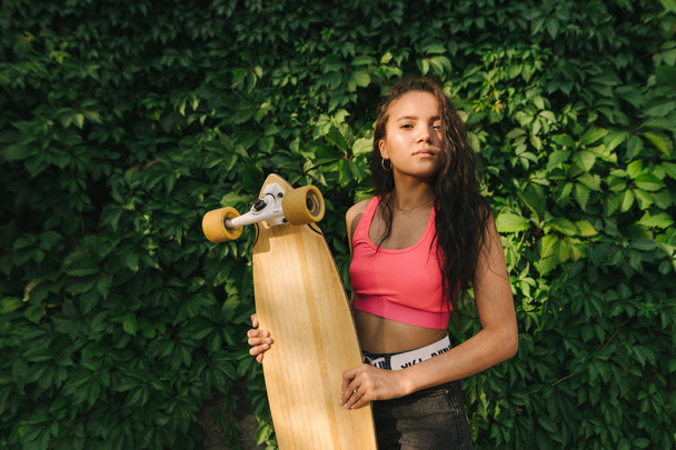 Closeup portrait of a Hispanic girl standing with a longboard in her hands against a background of green wall with ivy, looking into the camera with a serious face, wearing stylish street clothes - Photo, Image