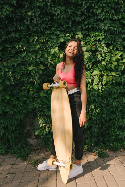 Smiling mulatto girl in stylish street clothes stands with a longboard in her hand on a background of a green wall covered with a vine, looks into the camera and smiles. Skateboarding concept - Foto, imagen
