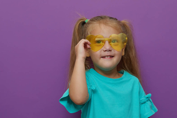 Portrait of little toddler child girl wearing light clothes and glasses isolated on purple background. Holds hands near the face. Happy childhood, wow and shock concept - Φωτογραφία, εικόνα