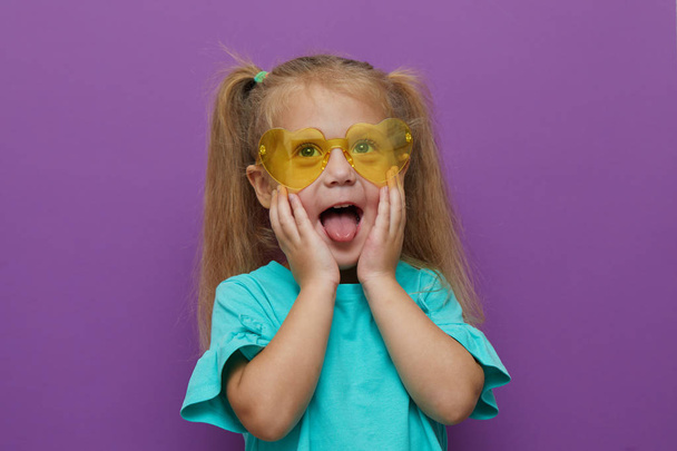 Portrait of little toddler child girl wearing light clothes and glasses isolated on purple background. Holds hands near the face. Happy childhood, wow and shock concept - Fotoğraf, Görsel