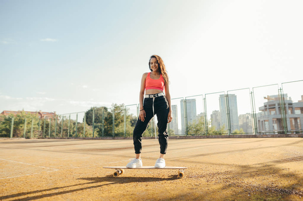 Ευτυχισμένο ισπανόφωνο κορίτσι στέκεται σε ένα skateboard στην παιδική χαρά, κοιτάζει κάμερα και χαμόγελα, φοράει κομψά ρούχα. Χαμογελαστό κορίτσι βόλτες στο longboard στο παλιό γήπεδο τέννις. Σκέιτ έννοια. Πολιτισμός του δρόμου. - Φωτογραφία, εικόνα