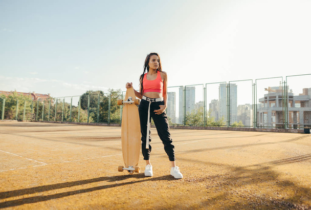Attractive girl in trendy street clothes stands with a longboard in hand on the playground, posing at the camera,looking away. Hispanic girl model with skateboard in hands stands on old tennis court. - Photo, Image