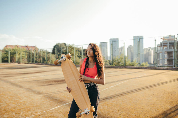 Street portrait of a mulatto stands on the playground,holds a longboard in hands, looks into the camera, wears stylish casual clothes. Curly hispanic girl model posing with a longboard in her hands. - Photo, Image