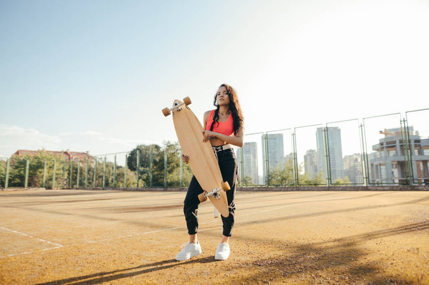 Mooi krullend mulatto meisje in casual kleding staat met een longboard in handen op een tennisbaan, kijkt in de camera en poses.Hispanic meisje met skateboard in handen poseren op speeltuin.Street cultuur. - Foto, afbeelding