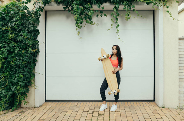 Beautiful young girl stands with longboard in hands on background of garage door with ivy, looks into camera, wears stylish street clothes. Stylish girl with a skateboard stands on a white background - Photo, Image