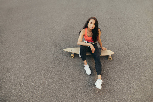 Πορτρέτο του ελκυστικό κορίτσι κάθεται στην άσφαλτο με longboard, κοιτάζοντας κάμερα με σοβαρό πρόσωπο, φορώντας κομψά casual ρούχα. Ισπανόφωνη κοπέλα με skateboard απομονωμένη σε άσφαλτο φόντο - Φωτογραφία, εικόνα