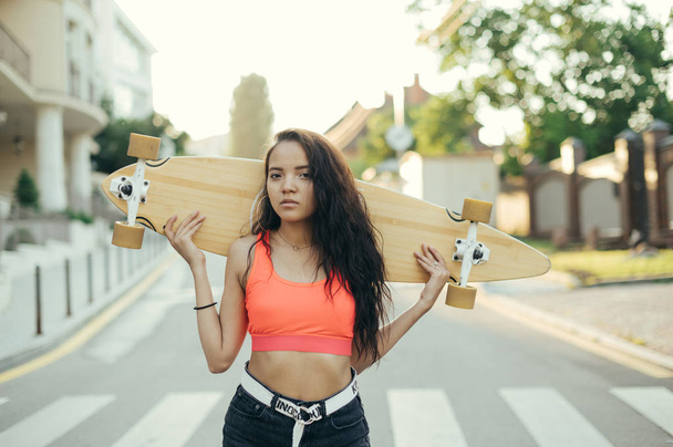Retrato de cerca de una atractiva chica hispana de pie con longboard en sus manos sobre un fondo de paisaje callejero al atardecer, mirando en cámara.
 - Foto, imagen
