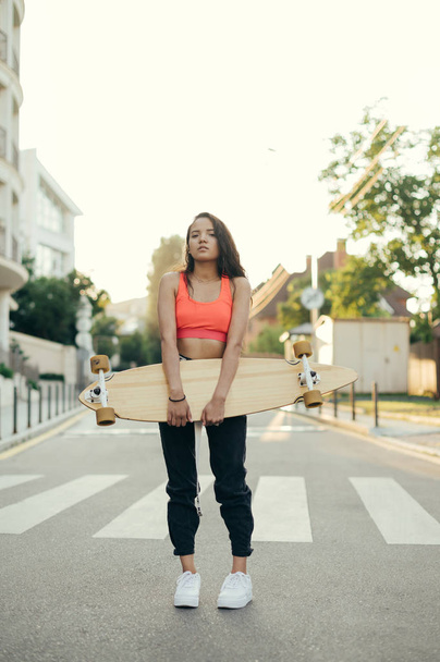 Portrait of stylish rider girl in trendy casual clothes with logboard in hands stands on street background at sunset and look at camera. Evening street portrait of girl with blackboard. Vertical - Foto, Imagem