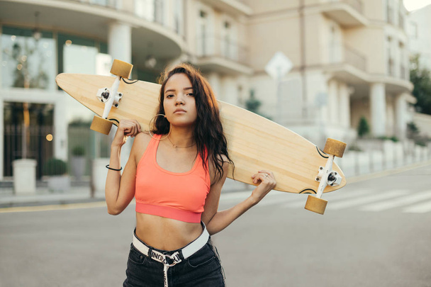 Menina piloto com skate na mão fica na rua. Retrato de rua de uma mulata segurando um longboard em suas mãos, olhando para a câmera, andando com uma placa à noite
. - Foto, Imagem