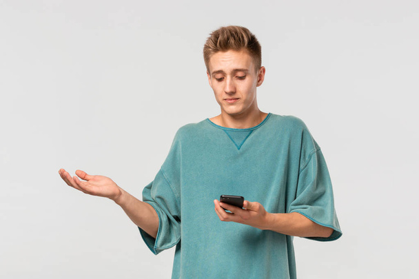 Confused blond teenager in a green tee looking at the screen of a smartphone isolated over light grey background. - Fotoğraf, Görsel