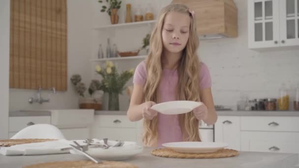 Young blond Caucasian girl in pink dress serving the table and smiling. Diligent teenager doing household work at the modern kitchen - Séquence, vidéo