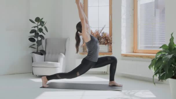 Young fit sporty brunette girl with pigtail in gray top, black leggings does exercises on mat in bright room. Doing sports and fitness training at home. Healthy lifestyle. stretching, leaning forward - Video, Çekim