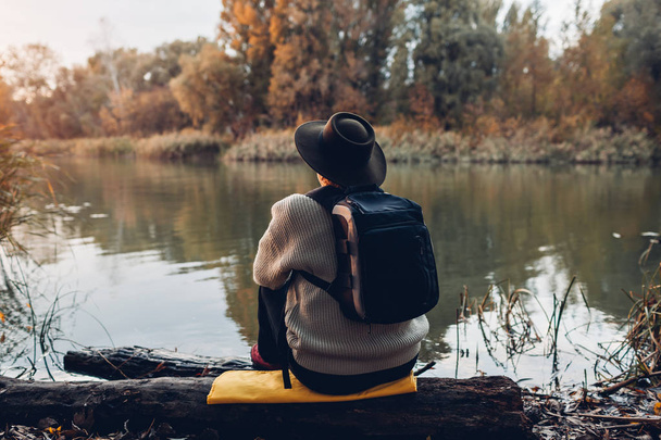 Viajero con mochila sentado en la orilla del río al atardecer. Mujer de mediana edad admirando la naturaleza otoñal
 - Foto, Imagen