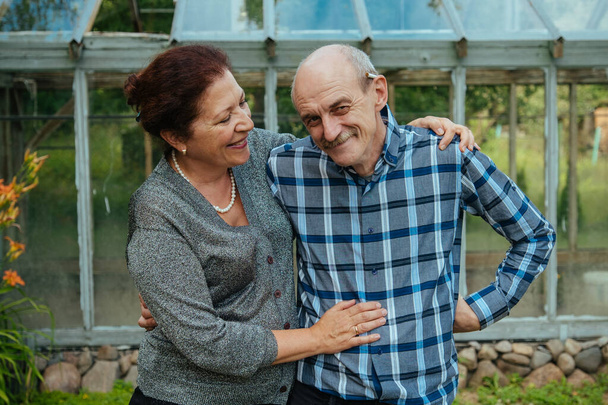 Smiling lovely old happy couple in front a glass greenhouse near summer house - Foto, Imagen