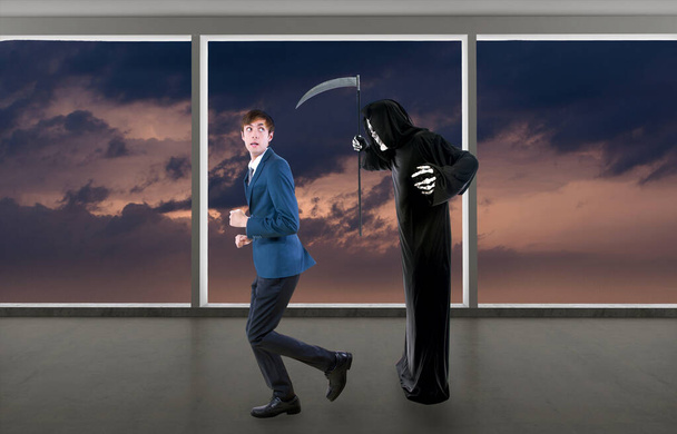 Man in a Halloween grim reaper ghost costume chasing, mocking and making fun of scared businessman running away.  Can also depict death following a man as a metaphor for life insurance.  - Photo, Image