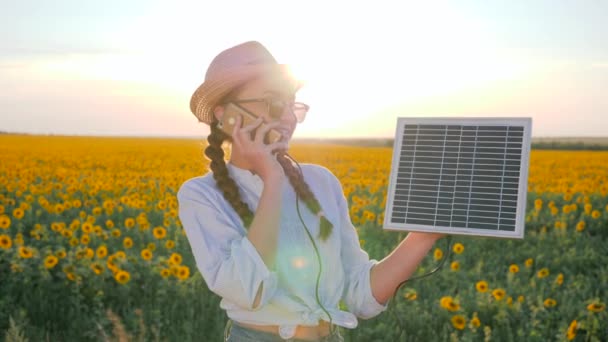 girl speaks by mobile phone and holds solar panel in background field of sunflowers, young woman with phone and solar battery - Footage, Video