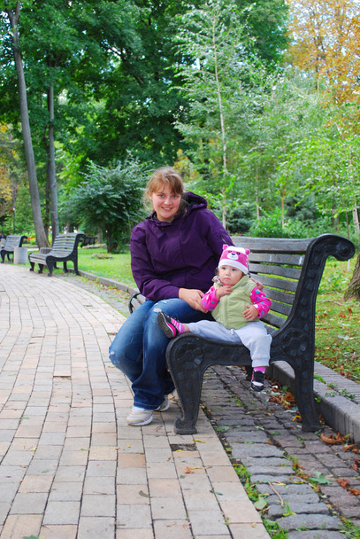 En el parque, mamá sentada en el banco y sosteniendo a un bebé daughte
 - Foto, Imagen
