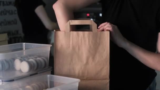 Κορίτσι ζαχαροπλαστικής με μαύρη στολή βάζει κουτιά με φρέσκα αμυγδαλωτά σε μια μεγάλη χάρτινη σακούλα. Προσεκτική συσκευασία των cookies για την παράδοση στους πελάτες. Τούρτες διακοπών από το αγαπημένο σας ζαχαροπλαστείο. - Πλάνα, βίντεο
