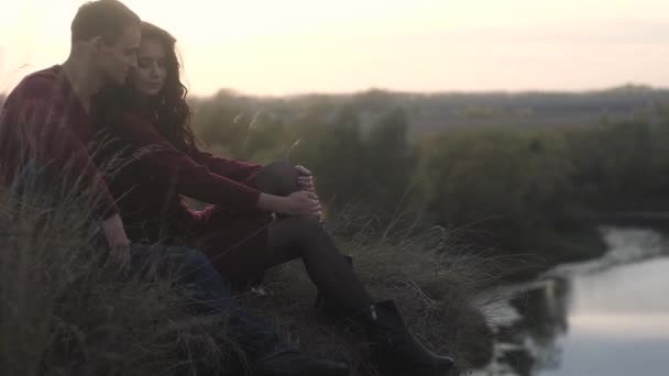 man and a beautiful girl in a dark dress with black loose hair sitting on a cliff near the river. They embrace and look into the distance. Autumn romantic mood on the background of the evening sunset. - Footage, Video