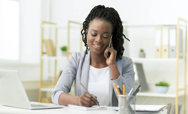 Mujer sonriente hablando por celular tomando notas sentada en el lugar de trabajo
 - Foto, imagen