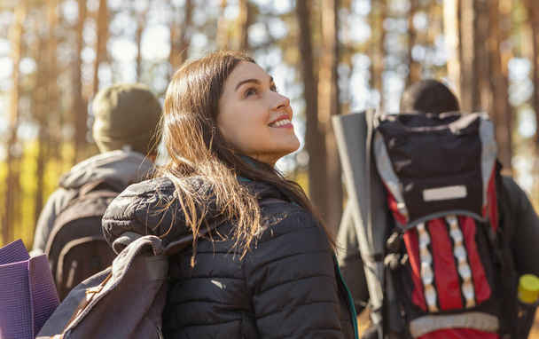 3人の観光客が秋の森をハイキングし、女の子は周りを見る - 写真・画像