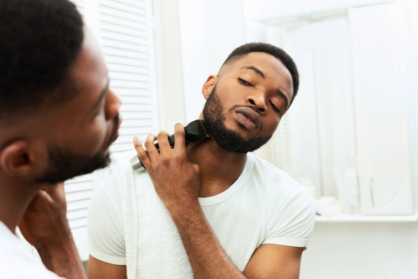 Young black man looking at mirror and shaving beard with trimmer - Fotoğraf, Görsel