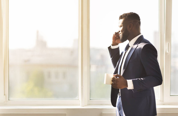 Atractivo hombre de negocios hablando en el teléfono inteligente y beber café cerca de la ventana
 - Foto, Imagen