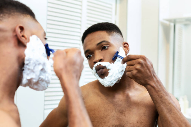 Африканский американец бреется в ванной, смотрит в зеркало
 - Фото, изображение