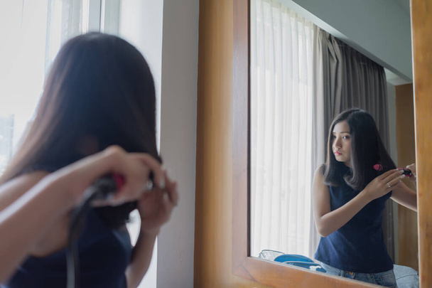 Молодая азиатка выпрямляет волосы выпрямителем плоского железа, глядя в зеркало дома
. - Фото, изображение
