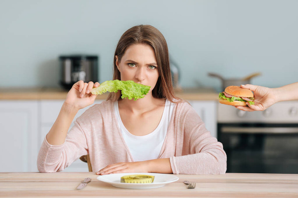 Sad woman eating healthy lettuce and female hand offering her a tasty burger. Diet concept - Φωτογραφία, εικόνα