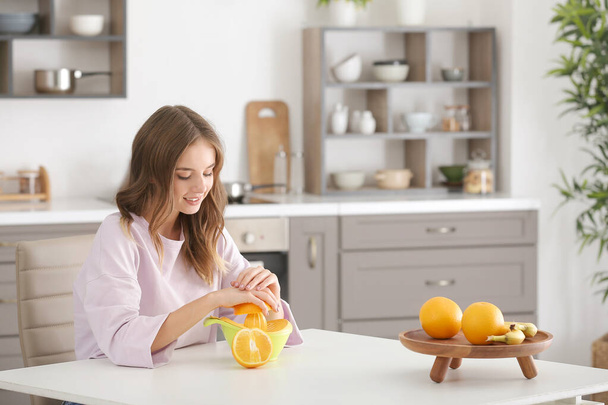 Όμορφη νεαρή γυναίκα κάνει φρέσκο χυμό πορτοκαλιού στην κουζίνα - Φωτογραφία, εικόνα