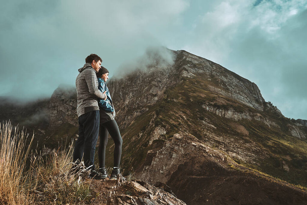 a young, happy couple hugging and standing on a cliff in the mountains looking forward - Foto, Imagem