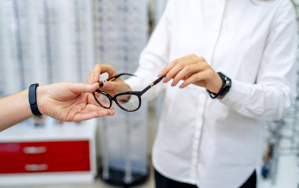 Row of glasses at an opticians. Eyeglasses shop. Stand with glasses in the store of optics. Woman's hand gives glasses to a man. Presenting spectacles. Closeup - Foto, afbeelding