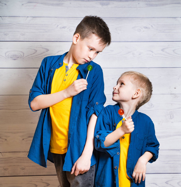 Portrait of two beautiful boys in yellow T-shirts and denim jackets, shirts. Boys are standing on a white wooden background. Little brothers. Cute boys with lollipops look at each other. Closeup. - Photo, Image