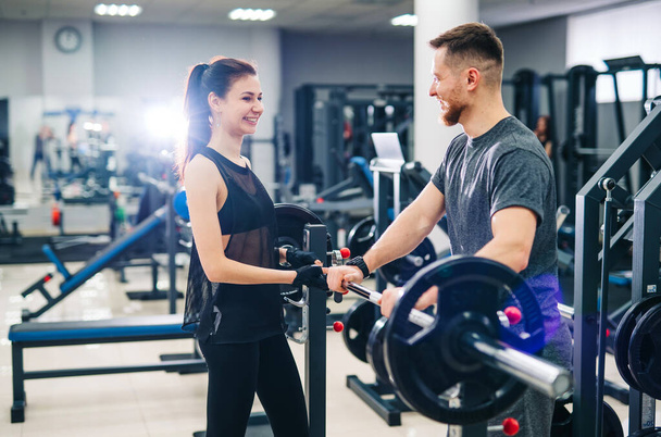 Sport, bodybuilding, lifestyle and people concept - young man and woman with a barbell. Trainer and fitness girl are having workout at gym. - Photo, image