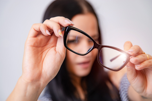 Portrait of a young woman with spectacles in hands. Blurred white background. Girl looks through eyeglasses. Longhaired brunette beautiful girl and black framed eyeglasses. Closeup. - Φωτογραφία, εικόνα