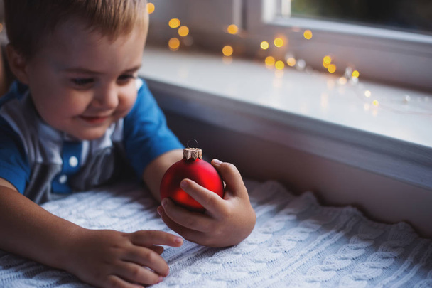 Niño feliz sosteniendo bola roja de Navidad cerca de la ventana interior con luces de guirnalda cálidas sobre fondo borroso
. - Foto, imagen