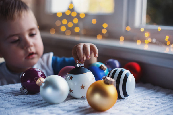 Ευτυχισμένο παιδί αγόρι επιλέγοντας μπάλα Χριστουγέννων κοντά στο παράθυρο εσωτερικό με ζεστά φώτα γιρλάντα σε θολή φόντο. - Φωτογραφία, εικόνα