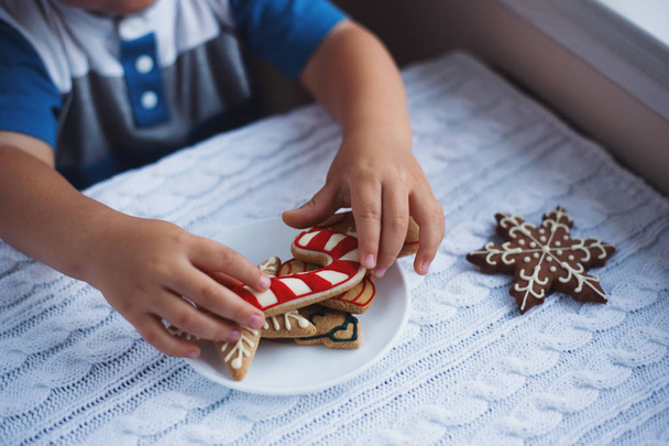 Παιδί αγόρι λαμβάνοντας τα Χριστούγεννα μελόψωμο από το πιάτο στο τραπέζι κοντά στο παράθυρο στο φως της ημέρας. - Φωτογραφία, εικόνα