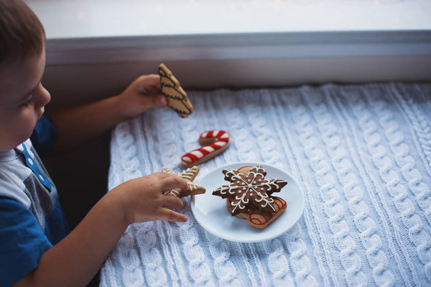Child boy taking Christmas gingerbread from plate on table near window in daylight. - Photo, Image